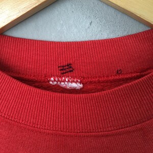 May include: Close-up of a red sweatshirt collar with ribbed knit texture. The collar has a small, torn tag with illegible text and a faded black graphic. The sweatshirt is hanging on a wooden hanger.