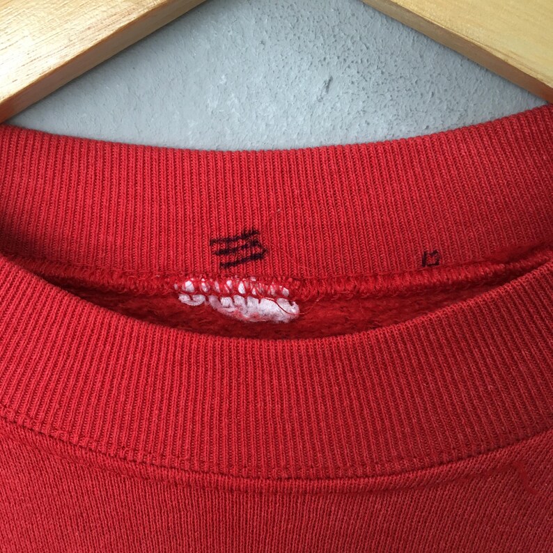 May include: Close-up of a red sweatshirt collar with ribbed knit texture. The collar has a small, torn tag with illegible text and a faded black graphic. The sweatshirt is hanging on a wooden hanger.