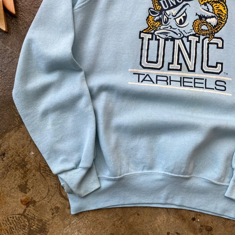 May include: A light blue crewneck sweatshirt featuring a graphic of a ram mascot wearing a hat above the text UNC and TARHEELS in navy blue and white lettering. The garment has long sleeves with ribbed cuffs and a ribbed hem at the bottom edge.