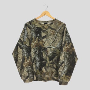 Puede incluir: Una sudadera de manga larga con un patr&oacute;n de camuflaje. El dise&ntilde;o presenta una mezcla de colores marr&oacute;n, verde y beige, imitando un entorno forestal. La sudadera est&aacute; colgada en una percha de madera.