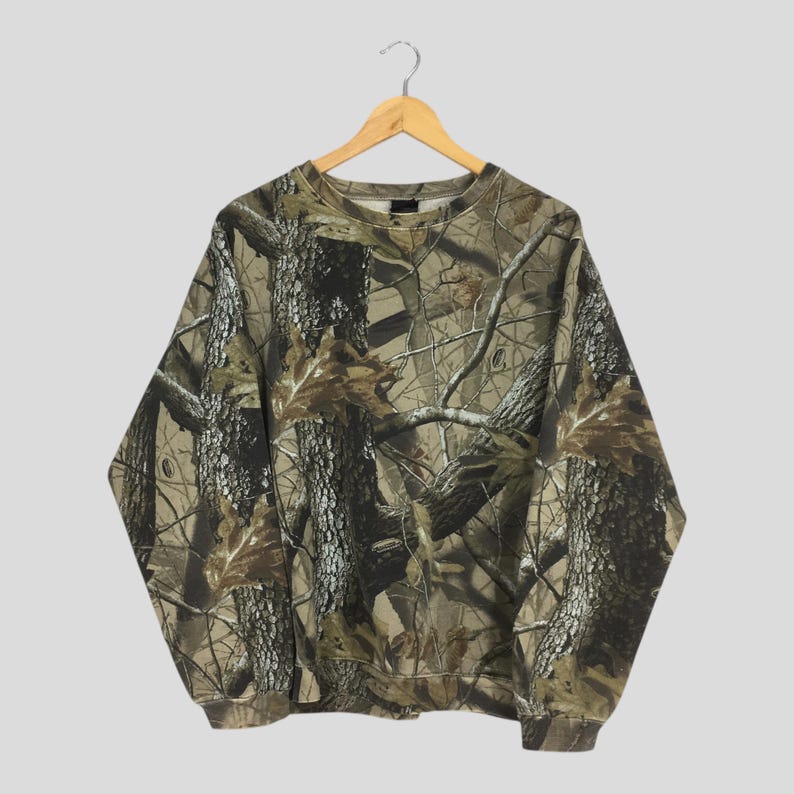 Puede incluir: Una sudadera de manga larga con un patr&oacute;n de camuflaje. El dise&ntilde;o presenta una mezcla de colores marr&oacute;n, verde y beige, imitando un entorno forestal. La sudadera est&aacute; colgada en una percha de madera.