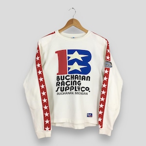 May include: Cream-colored long-sleeve shirt with red star stripes on the sleeves. The front features a large blue letter "B" with two white stars, and the text "BUCHANAN RACING SUPPLYCO" in black. The text "BUCHANAN, MICHIGAN" is also printed on the shirt.