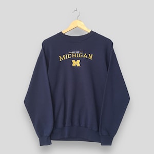 May include: A navy blue long-sleeve crewneck sweatshirt featuring the word MICHIGAN in yellow arched text above a yellow block letter M and the text EST. 1817. The garment is displayed on a light-coloured wooden hanger against a plain background.