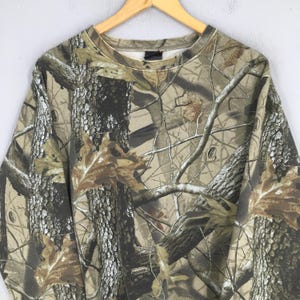 Puede incluir: Una sudadera con estampado de camuflaje con cuello redondo, colgada de una percha de madera. El dise&ntilde;o presenta una mezcla de colores marr&oacute;n, verde y beige, imitando un entorno forestal. La tela parece ser de peso medio.