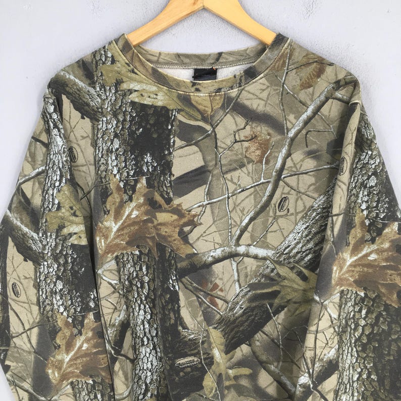 Puede incluir: Una sudadera con estampado de camuflaje con cuello redondo, colgada de una percha de madera. El dise&ntilde;o presenta una mezcla de colores marr&oacute;n, verde y beige, imitando un entorno forestal. La tela parece ser de peso medio.
