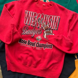 May include: A red sweatshirt with the text "WISCONSIN Badgers Rose Bowl Champions January 1, 1994" in white and black. The sweatshirt has a crew neck and long sleeves. Other sweatshirts in green, black, and yellow are visible.