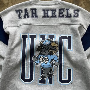Puede incluir: Sudadera gris con "TAR HEELS" impreso en los hombros. La parte delantera presenta un gran gr&aacute;fico "UNC" con una mascota de carnero con una camiseta azul claro. Detalles en azul marino en las mangas.