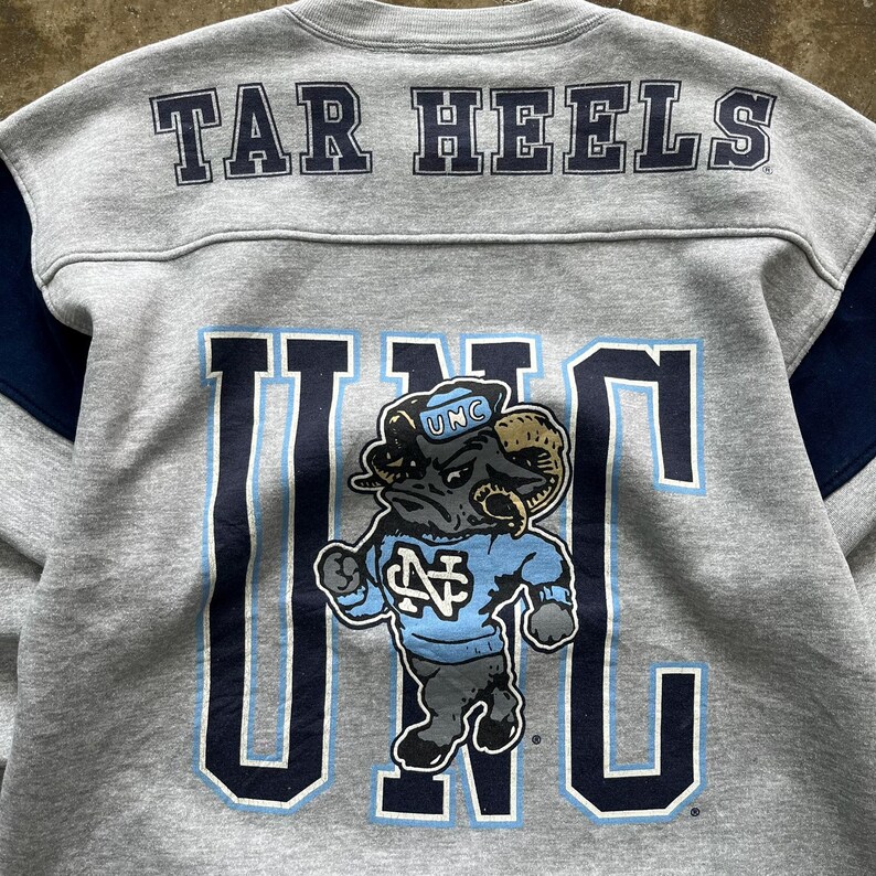 Puede incluir: Sudadera gris con "TAR HEELS" impreso en los hombros. La parte delantera presenta un gran gr&aacute;fico "UNC" con una mascota de carnero con una camiseta azul claro. Detalles en azul marino en las mangas.