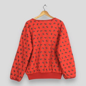 May include: A red sweatshirt with a pattern of small, repeating black and yellow figures. The sweatshirt has a crew neck and long sleeves, with red ribbing at the cuffs and hem. The garment is hanging on a wooden hanger.