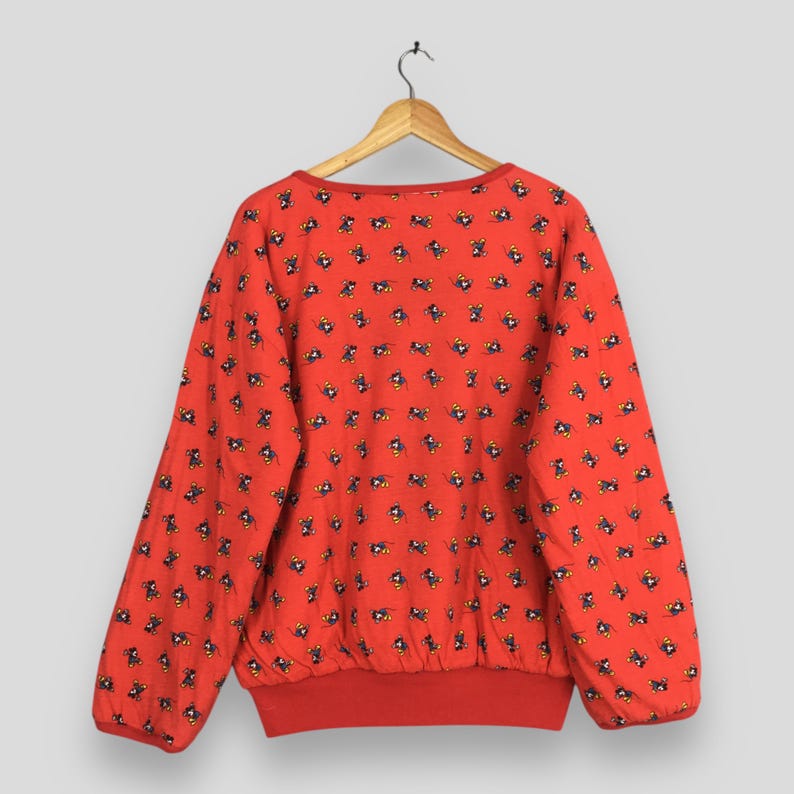 May include: A red sweatshirt with a pattern of small, repeating black and yellow figures. The sweatshirt has a crew neck and long sleeves, with red ribbing at the cuffs and hem. The garment is hanging on a wooden hanger.