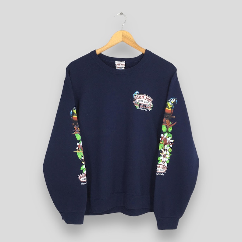 Puede incluir: Sudadera azul marino con cuello redondo. Las mangas presentan un dise&ntilde;o colorido de loros, flores y el texto "Ron Jon Surf Shop". Una versi&oacute;n m&aacute;s peque&ntilde;a del logotipo est&aacute; en la parte superior del pecho. La sudadera est&aacute; colgada de una percha de madera.