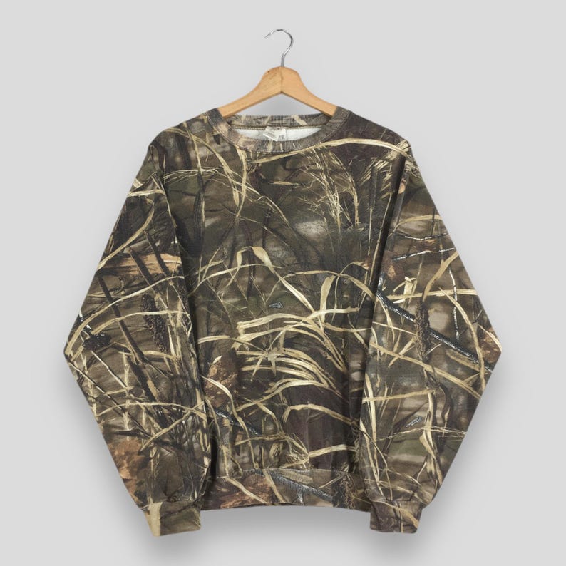 May include: A camouflage-patterned sweatshirt hangs on a wooden hanger. The long-sleeved top features a blend of brown, green, and tan hues, mimicking a natural, grassy environment. The sweatshirt has a crew neck and ribbed cuffs and hem.