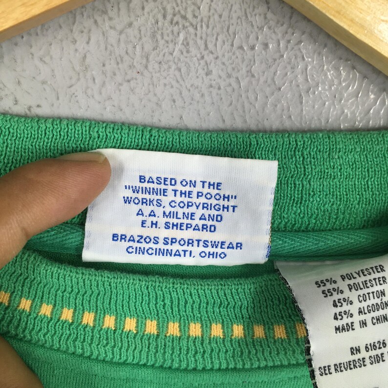 May include: Close-up of a green shirt with a tag that reads "BASED ON THE 'WINNIE THE POOH' WORKS, COPYRIGHT A.A. MILNE AND E.H. SHEPARD BRAZOS SPORTSWEAR CINCINNATI, OHIO." The shirt also has a yellow and white design.