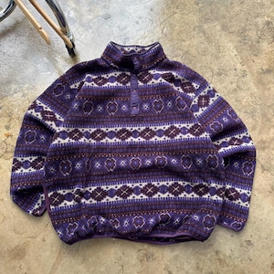 May include: A purple fleece pullover with a high collar and button closure. The sweater features a geometric pattern in white and dark purple. The fleece has a relaxed fit and a drawstring hem. A casual, warm garment for cooler weather.