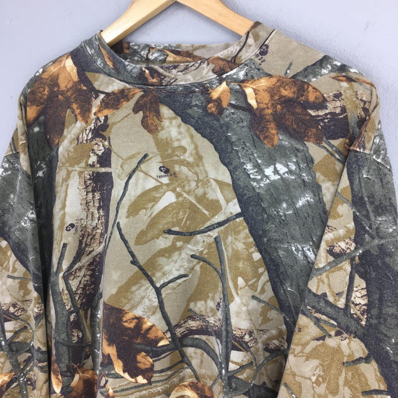 May include: A long-sleeved camouflage shirt in shades of brown, green, and tan, featuring a tree and leaf pattern. The shirt is hanging on a wooden hanger. The fabric has a soft appearance.