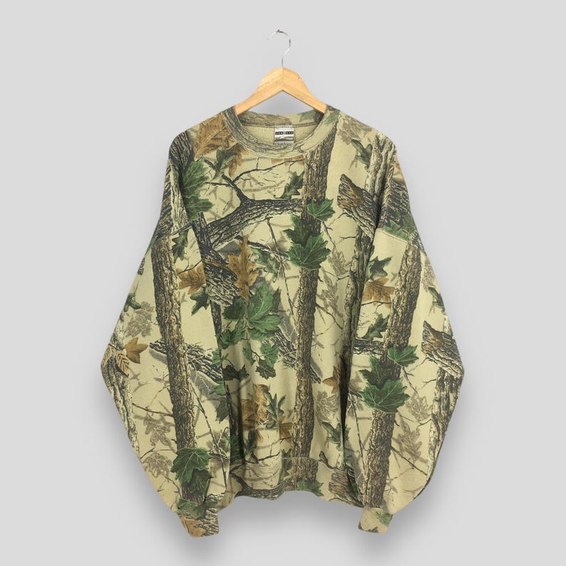 May include: A camouflage-patterned sweatshirt with a crew neck, featuring a design of tree branches and green and brown leaves. The long-sleeved top is hanging on a wooden hanger.