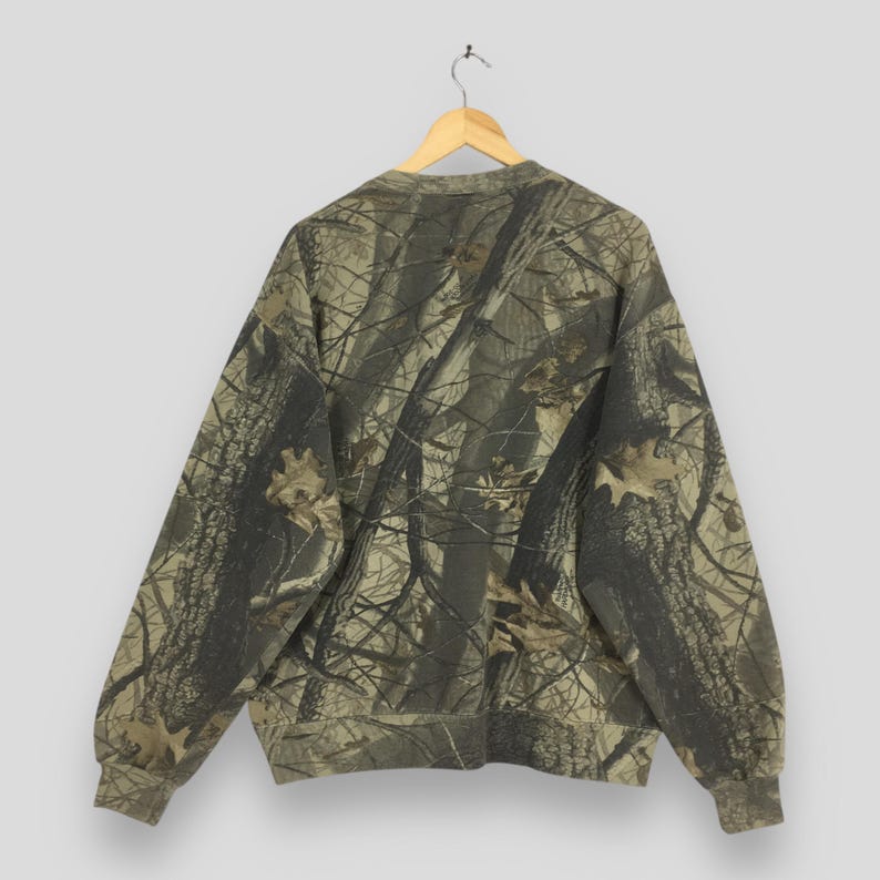 May include: A camouflage-patterned sweatshirt hangs on a wooden hanger. The long-sleeved top features a repeating design of tree branches and leaves in shades of brown, green, and tan. The sweatshirt is made of a soft, comfortable material.