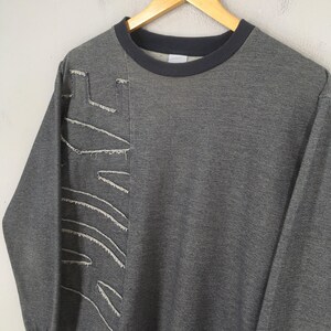 May include: A gray sweatshirt with a dark gray crew neck and cuffs. The left side features a distressed, stitched design in white thread. The sweatshirt is hanging on a wooden hanger against a neutral background.