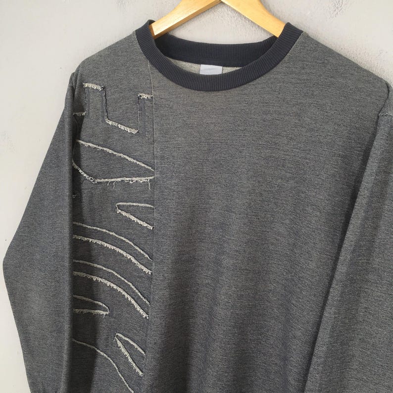May include: A gray sweatshirt with a dark gray crew neck and cuffs. The left side features a distressed, stitched design in white thread. The sweatshirt is hanging on a wooden hanger against a neutral background.