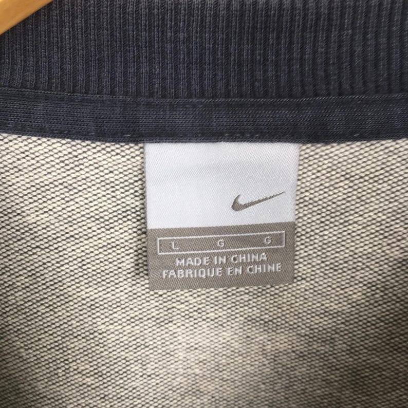 May include: Close-up of a gray and navy blue garment tag. The tag features the Nike swoosh logo and the text "MADE IN CHINA". The garment appears to be a sweatshirt or similar apparel item.