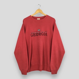 Op de afbeelding: Een rode crewneck sweatshirt met de tekst "UNIVERSITY OF GEORGIA" in zwart en rood. De sweatshirt hangt aan een houten hanger tegen een lichte achtergrond. Het heeft lange mouwen.