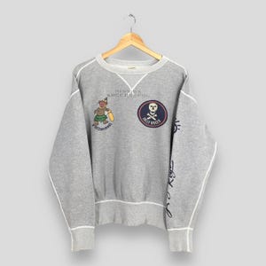 May include: A gray sweatshirt with white trim and a crew neck. The sweatshirt features a Jolly Roger patch, a cartoon character, and the words "Mission Successful" and "Asterperious". The sleeve has cursive text.