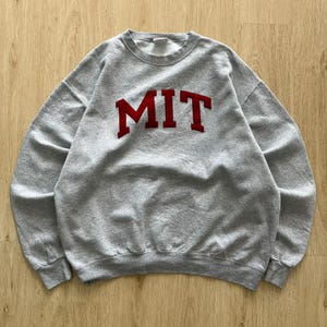Könnte beinhalten: Ein graues Sweatshirt mit Rundhalsausschnitt und den roten Buchstaben "MIT" auf der Brust. Das Sweatshirt besteht aus einem weichen Material und hat einen gerippten Ausschnitt, Bündchen und Saum. Ein klassischer Kleidungsstil.