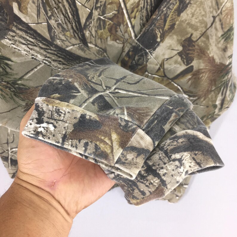 May include: A camouflage-patterned long-sleeve shirt. The fabric features a mix of brown, green, and tan hues, mimicking a forest environment. The cuffs are folded over, revealing the interior fabric. The shirt is made of a soft, comfortable material.