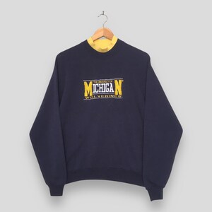 May include: A navy blue long-sleeve crewneck sweatshirt featuring a yellow and white embroidered graphic on the chest that reads THE UNIVERSITY OF MICHIGAN WOLVERINES. The garment has a yellow mock neck collar visible inside the neckline.