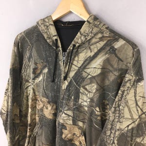 May include: A camouflage-patterned zip-up hoodie with a hood and a front pocket. The hoodie features a mix of brown, green, and tan colors, creating a realistic forest-like design. The zipper is black, and the interior is black.