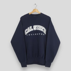Könnte beinhalten: Marineblaues Sweatshirt mit weißem Aufdruck "CAL STATE FULLERTON" in Bogenform. Das Kleidungsstück hängt an einem Holzbügel. Das Design deutet auf ein Universitäts- oder College-Thema mit entspannter Passform hin.