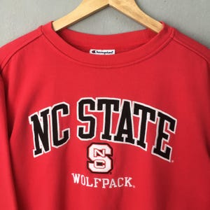 May include: A red Champion sweatshirt with the words "NC STATE" in white block letters with black trim. Below the text is a white "S" with "NC" inside and the word "WOLFPACK" in white.