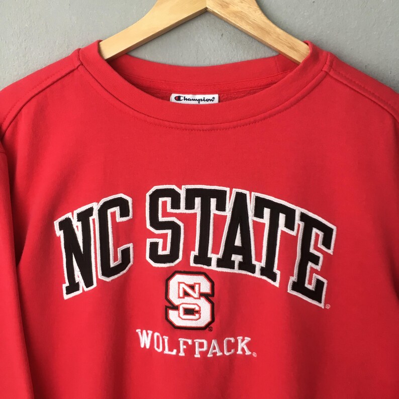 May include: A red Champion sweatshirt with the words "NC STATE" in white block letters with black trim. Below the text is a white "S" with "NC" inside and the word "WOLFPACK" in white.