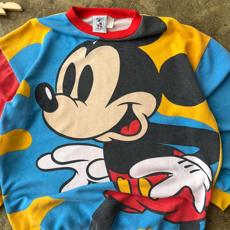 K&ouml;nnte beinhalten: Ein Vintage-Sweatshirt mit Rundhalsausschnitt und einer gro&szlig;en Mickey Mouse-Grafik. Das Sweatshirt hat ein Colorblock-Design mit blauen, gelben, roten und grauen Eins&auml;tzen. Der Kragen ist rot und die &Auml;rmel sind lang.