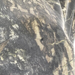 Puede incluir: Primer plano de una tela con estampado de camuflaje en tonos verdes, marrones y grises. La tela tiene un aspecto moteado con una peque&ntilde;a mancha oscura. El patr&oacute;n recuerda a un bosque o entorno boscoso.