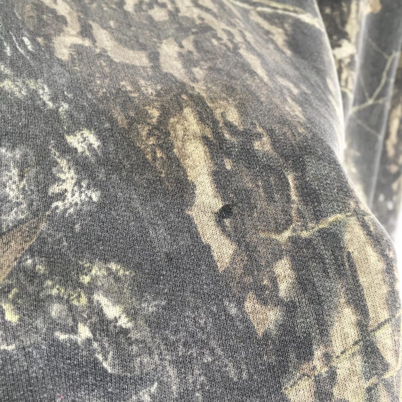 Puede incluir: Primer plano de una tela con estampado de camuflaje en tonos verdes, marrones y grises. La tela tiene un aspecto moteado con una peque&ntilde;a mancha oscura. El patr&oacute;n recuerda a un bosque o entorno boscoso.