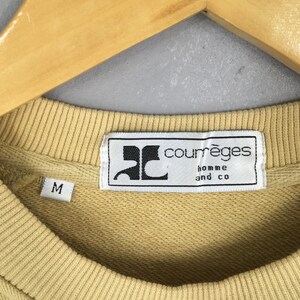 May include: Close-up of a light yellow sweatshirt with a crew neck. The label reads "courr&egrave;ges homme and co" in black text on a white background. The size "M" is visible on the left side of the sweatshirt.