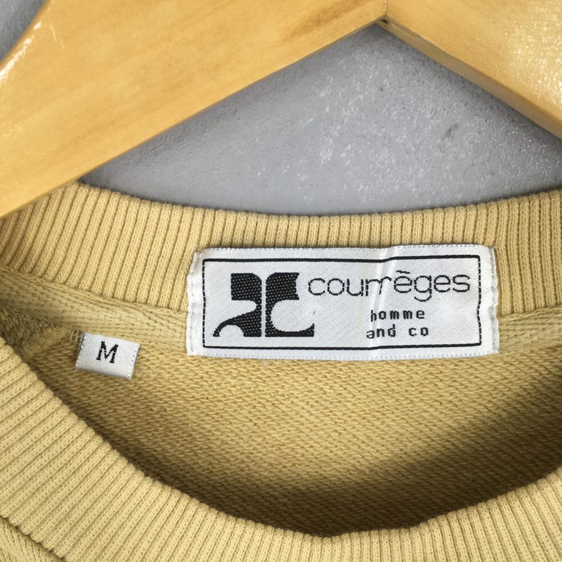 May include: Close-up of a light yellow sweatshirt with a crew neck. The label reads "courr&egrave;ges homme and co" in black text on a white background. The size "M" is visible on the left side of the sweatshirt.