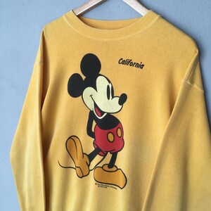 May include: Mustard yellow sweatshirt featuring a large graphic of Mickey Mouse in his classic pose, with red shorts and yellow shoes. The word "California" is printed above the character. The sweatshirt is hanging on a wooden hanger.