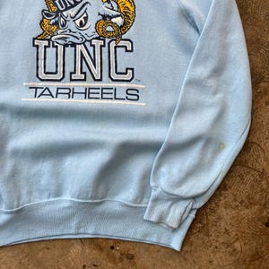 May include: A light blue long-sleeve sweatshirt featuring a graphic of a ram mascot wearing a cap, the text UNC in large block letters, and the word TARHEELS underneath. The design is printed in navy blue, white, and yellow on the chest.