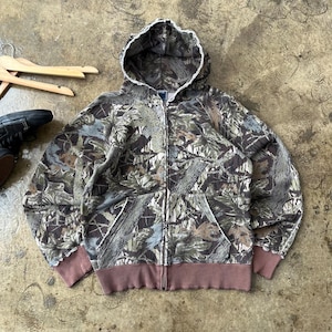 K&ouml;nnte beinhalten: Ein Kapuzenpullover mit Rei&szlig;verschluss und Camouflage-Muster. Die Jacke hat ein braunes, gr&uuml;nes und graues Blattmuster, eine wei&szlig; gef&uuml;tterte Kapuze sowie einen braunen gerippten Saum und B&uuml;ndchen. Sie hat zwei Vordertaschen und einen Rei&szlig;verschluss.