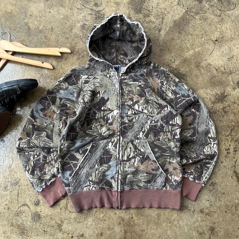 K&ouml;nnte beinhalten: Ein Kapuzenpullover mit Rei&szlig;verschluss und Camouflage-Muster. Die Jacke hat ein braunes, gr&uuml;nes und graues Blattmuster, eine wei&szlig; gef&uuml;tterte Kapuze sowie einen braunen gerippten Saum und B&uuml;ndchen. Sie hat zwei Vordertaschen und einen Rei&szlig;verschluss.