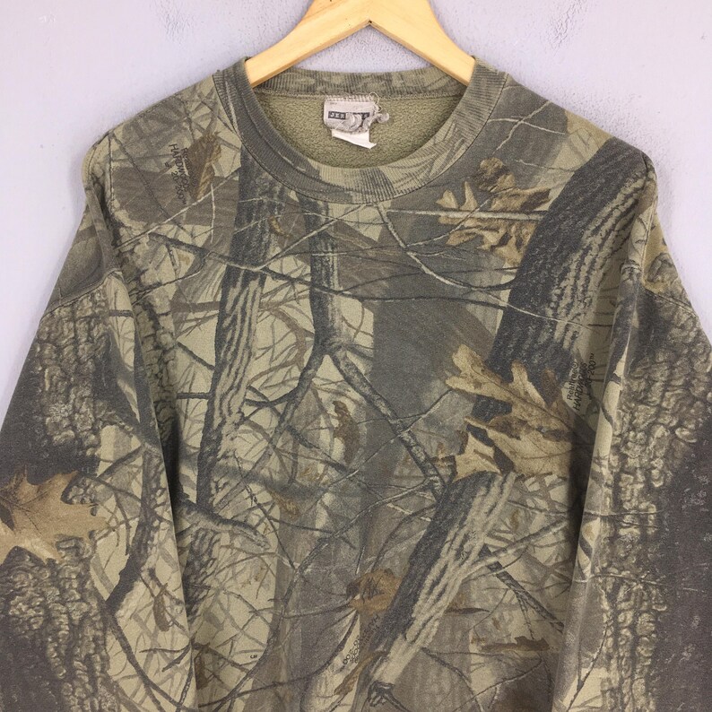 May include: A camouflage-patterned sweatshirt in shades of brown, green, and tan, hanging on a wooden hanger. The design mimics a forest environment, with tree branch and leaf details. The sweatshirt has a crew neck and long sleeves.