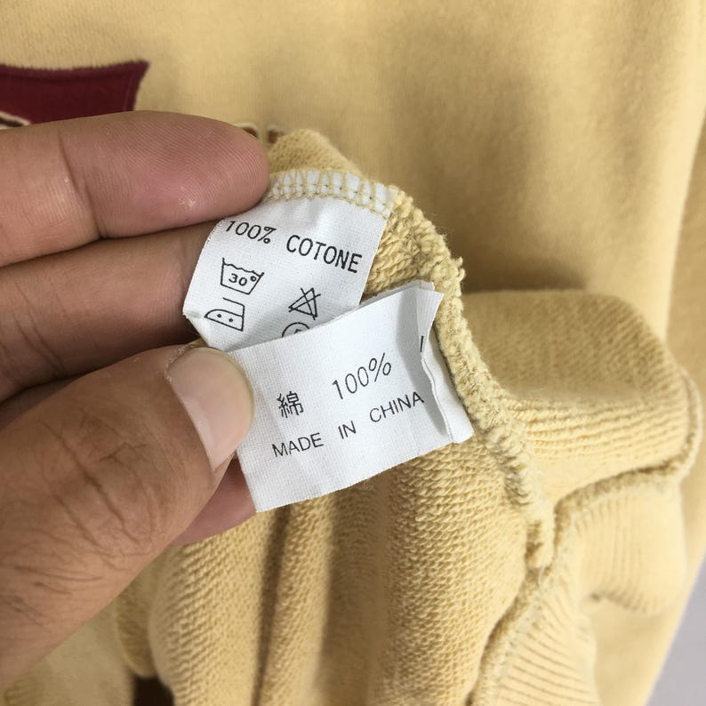 May include: Close-up of a light yellow sweatshirt with a tag that reads "100% COTONE" and "100%" in Japanese characters. The tag also includes washing instructions and "MADE IN CHINA".