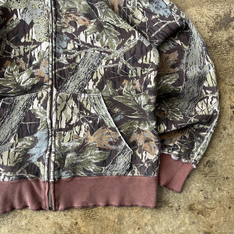 K&ouml;nnte beinhalten: Ein Kapuzenpullover mit Rei&szlig;verschluss und einem Tarnmuster in Braun-, Gr&uuml;n- und Graut&ouml;nen. Der Sweatshirt hat einen braunen gerippten Saum und B&uuml;ndchen sowie eine Vordertasche. Der Rei&szlig;verschluss ist silberfarben.