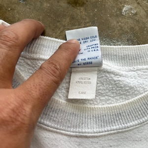 May include: A white sweatshirt with a ribbed crew neckline and two interior clothing labels. The top label reads "SAVE THE RHINOS" and "CRAFTED WITH PRIDE IN U.S.A." The bottom label indicates a fabric composition of 60% cotton and 40% polyester, size XLARGE.