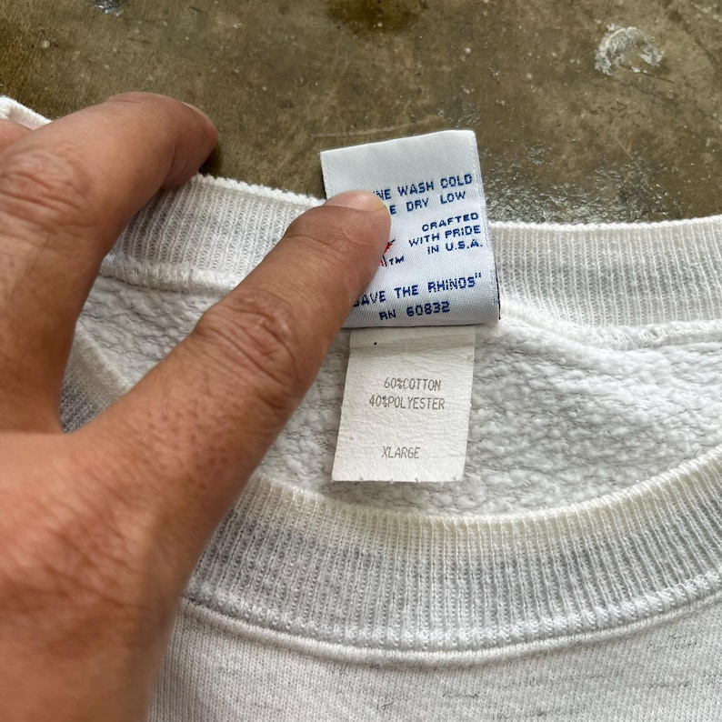May include: A white sweatshirt with a ribbed crew neckline and two interior clothing labels. The top label reads "SAVE THE RHINOS" and "CRAFTED WITH PRIDE IN U.S.A." The bottom label indicates a fabric composition of 60% cotton and 40% polyester, size XLARGE.