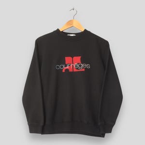 May include: Black crew neck sweatshirt with the Courr&egrave;ges logo in red and white. The logo features the brand name in a cursive font and a stylized red design. The sweatshirt is hanging on a wooden hanger.