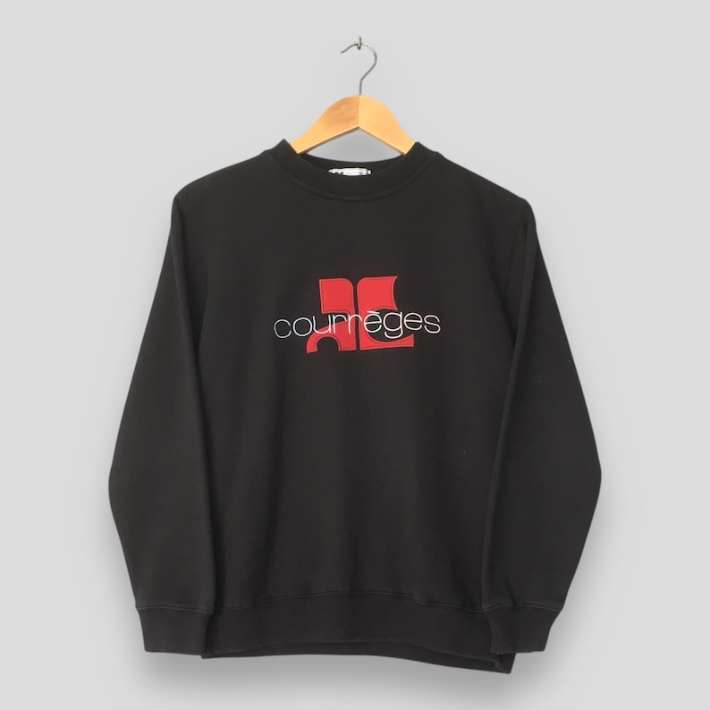 May include: Black crew neck sweatshirt with the Courr&egrave;ges logo in red and white. The logo features the brand name in a cursive font and a stylized red design. The sweatshirt is hanging on a wooden hanger.