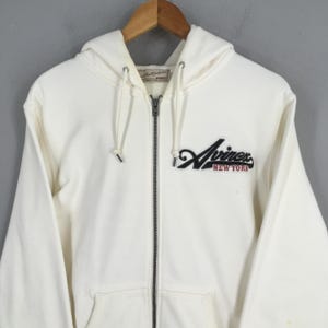 Puede incluir: Sudadera con capucha con cremallera de color crema con cord&oacute;n. La parte delantera presenta un logotipo bordado en negro y rojo que dice "Aviirex NEW YORK". La sudadera con capucha tiene una cremallera completa y dos bolsillos delanteros. La prenda est&aacute; hecha de un material suave y c&oacute;modo.